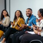 A group of diverse young professionals are laughing and smiling as they look at their phones during a conference or meeting. They are all wearing name badges and appear to be engaged in conversation.