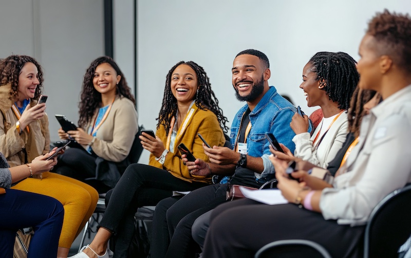 A group of diverse young professionals are laughing and smiling as they look at their phones during a conference or meeting. They are all wearing name badges and appear to be engaged in conversation.