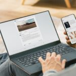 Young Man at Home is Using Laptop Computer, Srufing the Net, Watching News, while Holding Smartphone with Social Media Interface. He's Sitting On a Couch in His Cozy Living Room. Over Shoulder Shot