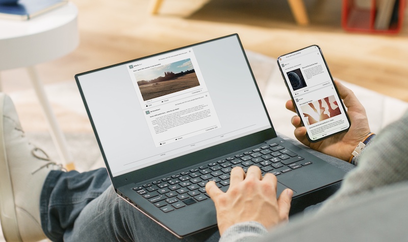 Young Man at Home is Using Laptop Computer, Srufing the Net, Watching News, while Holding Smartphone with Social Media Interface. He's Sitting On a Couch in His Cozy Living Room. Over Shoulder Shot
