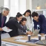 Colleagues discuss a business project by comparing analysis data and graphs on a laptop.