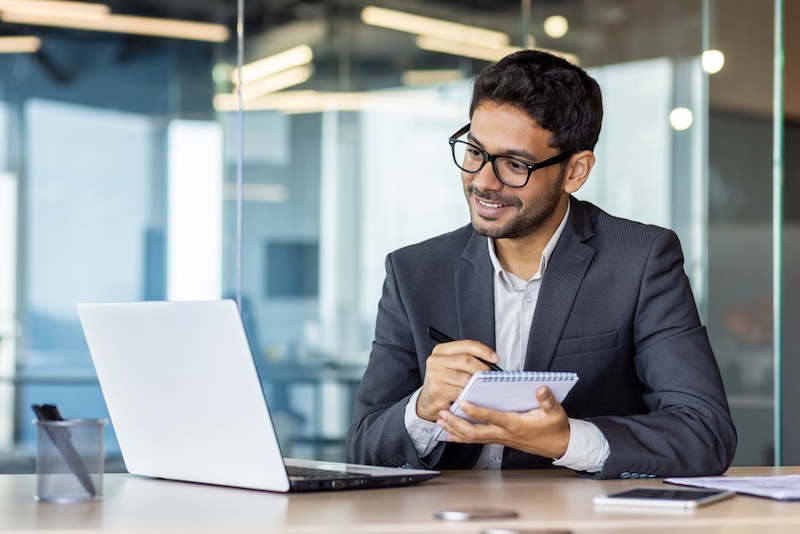 Mature experienced hispanic businessman in middle of office watching online webinar and training course, man using laptop for video call, writing data in notebook, smiling excitedly and satisfied
