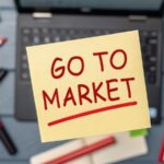 B2BMX GTM Strategy & Advanced ABM Track startup success execution competition goals concept. Sticky note with "Go to Market" on a desk with office supplies.