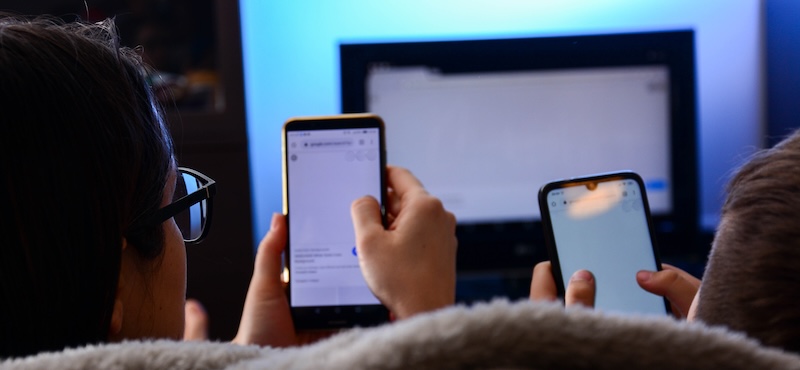 Connected TV Circana’s research friendship, people, technology and entertainment concept A mother and son holds a smartphones in theirs hands using a DLNA in the living room in front of the TV, enjoying numerous content over